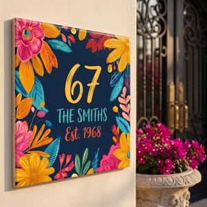 Puede incluir: Un letrero cuadrado para casa con fondo azul oscuro y un borde floral colorido en tonos rosa, amarillo y naranja. El letrero muestra el número 67 en una fuente amarilla grande, el texto THE SMITHS en turquesa y Est. 1968 en naranja.