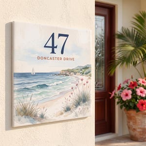 Könnte beinhalten: Ein quadratisches Hausschild mit einer Küstenszene im Aquarellstil, einem Segelboot auf blauem Wasser, einem Sandstrand und rosa Blumen. Der Text 47 ist in großer dunkelblauer Schrift über dem Text DONCASTER DRIVE in brauner Schrift gedruckt.