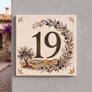 以下が含まれることがあります： ダークブラウンの数字「19」が描かれた正方形のセラミック製番地プレート。左側にはオリーブの木、下部には丘陵の風景、右側には緑と黒のオリーブの実がついた湾曲した枝が描かれています。