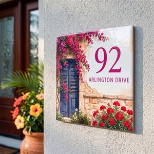 Peut inclure: Une plaque de numéro de maison rectangulaire affichant le nombre 92 et le texte ARLINGTON DRIVE. Le motif représente une scène peinte avec une porte bleue à volets, un mur en pierre, des bougainvilliers roses et des buissons de fleurs rouges.