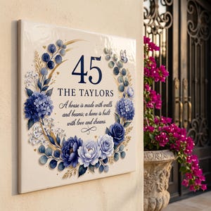 Puede incluir: Un letrero cuadrado de color crema para casa con una corona floral de hortensias azules, rosas y hojas de eucalipto. El texto dice 45, THE TAYLORS, y A house is made with walls and beams; a home is built with love and dreams.