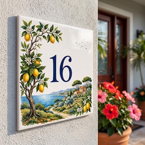 Könnte beinhalten: Ein quadratisches Hausnummernschild aus Keramik mit der Zahl 16 in Dunkelblau. Das Design zeigt links einen Zitronenbaum mit gelben Früchten und weißen Blüten sowie rechts unten eine Küstenlandschaft mit Häusern und dem Meer.