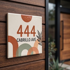 Peut inclure: Une plaque de numéro de maison carrée affichant le texte 444 CABRILLO AVE en lettres marron foncé. Le fond blanc cassé est orné de formes semi-circulaires superposées en terre cuite et vert sauge, ainsi qu'une illustration de feuille noire.
