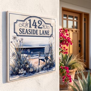 Puede incluir: Un letrero rectangular de cerámica para casa con el texto 142 SEASIDE LANE en letras azul oscuro. El diseño incluye una escena costera con una silla Adirondack azul, hierba de playa y detalles de estrellas de mar sobre un fondo crema.