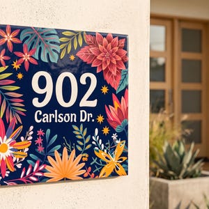 Könnte beinhalten: Ein quadratisches Hausnummernschild mit dunkelblauem Hintergrund, verziert mit bunten tropischen Blumen und Blättern. Der weiße Text zeigt die Nummer 902 und die Adresse Carlson Dr. Das Schild ist an einer hellen Außenwand montiert.