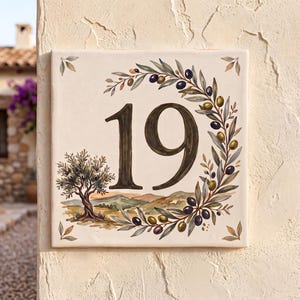 Peut inclure: Une plaque de numéro de maison carrée en céramique affichant le chiffre 19 en brun foncé. Le motif comprend une couronne de branches d'olivier peinte, un petit olivier et un paysage avec des collines dans des tons de vert, brun et crème.