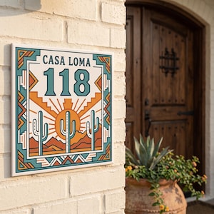 Puede incluir: Un letrero cuadrado de número de casa con el texto CASA LOMA y el número 118 en color verde azulado oscuro. El diseño incluye un paisaje desértico con tres cactus saguaro, un sol naciente y montañas en naranja y verde azulado.