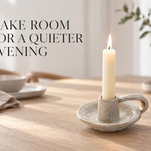 Puede incluir: Una vela cónica de color crema se encuentra en un candelabro de cerámica gris claro moteado con una pequeña asa. El texto "MAKE ROOM FOR A QUIETER EVENING" está impreso en negro a la izquierda de la imagen, sobre el candelabro.