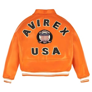 May include: A bright orange leather varsity jacket featuring the black text AVIREX arched across the upper back. Below the text is a circular patch with a shield graphic and the text ALL AMERICANS ESTABLISHED 1975. The word USA is in black.