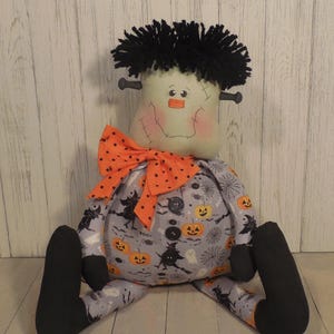 May include: A fabric doll featuring a light green head with black yarn hair, side bolts, and stitched facial features. The body is made of gray fabric with a pattern of orange pumpkins, black witches, and white ghosts, accented by three black buttons.