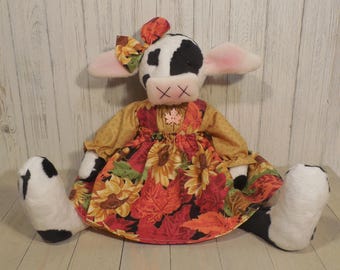 Könnte beinhalten: Eine Plüschkuh-Puppe mit schwarz-weißem Fell, bekleidet mit einem senfgelben Langarmshirt und einem Rock mit Sonnenblumen- und Herbstblattmuster. Die Puppe trägt eine passende Schleife am Kopf und ein gesticktes Ahornblatt auf der Brust.
