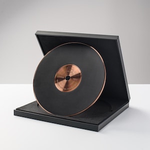 Può includere: Un tappetino circolare nero per giradischi con superficie testurizzata e un disco centrale in metallo color rame. Il disco riporta la scritta COPPER TRIO e ULTIMATE CONTROL PLATTER MAT. Il tappetino è esposto all'interno di una scatola nera aperta.