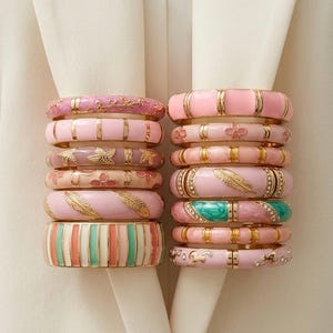 Peut inclure: Deux piles de bracelets rigides en métal doré avec de l'émail dans des tons rose, crème et bleu sarcelle. Les bracelets présentent divers motifs incluant des plumes dorées, des fleurs, des rayures et de petits cristaux incrustés.