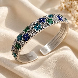 Peut inclure: Un bracelet manchette en argent orné d'un motif complexe de fleurs en relief avec un remplissage en émail bleu et vert. Le métal présente une finition oxydée pour souligner les détails floraux, et l'intérieur affiche des inscriptions gravées.