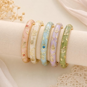 Pode incluir: Cinco pulseiras tipo bracelete em tons pastel de rosa, creme, azul claro, lavanda e verde. Cada pulseira apresenta um acabamento liso e brilhante com padrões florais e de folhas em tom dourado em relevo por toda a superfície.