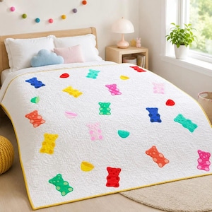 Può includere: Una trapunta bianca con un motivo ripetuto di forme di orsetti gommosi colorati in rosso, arancione, giallo, verde, blu e rosa. La trapunta presenta un bordo giallo e una superficie trapuntata, stesa su un letto con cuscini bianchi.
