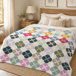 Op de afbeelding: Een witte quilt met een herhalend patroon van vierbladige bloemvormen in diverse kleuren, waaronder roze, geel, blauwgroen, donkergroen en lichtblauw. De quilt heeft een ruitvormig stiksel en ligt op een bed met beige kussens.
