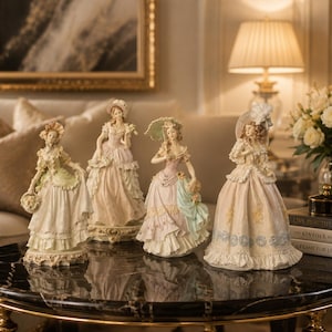 May include: Four porcelain figurines of individuals in elaborate historical gowns and dresses, each standing on a decorative base. The figures feature intricate lace details, pastel color palettes, and ornate hats, arranged on a dark marble surface.