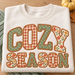 May include: A cream-colored crewneck sweatshirt featuring the text COZY SEASON in large block letters. The letters are filled with alternating patterns of green with white polka dots, orange and white gingham, and floral prints.