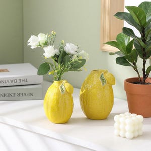 May include: Two bright yellow ceramic vases shaped like textured lemons, each featuring a small green leaf detail. One vase holds a bouquet of white artificial roses with green stems, while the other stands empty next to a white bubble candle.