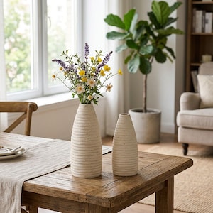 Op de afbeelding: Twee gebroken witte keramische vazen met een getextureerd, horizontaal geribbeld oppervlak. De langere vaas bevat een boeket van paarse lavendel en kleine gele en witte wilde bloemen. De vazen staan op een houten tafeloppervlak.