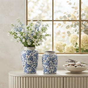 Op de afbeelding: Twee keramische vazen met een blauw-wit bloemmotief, waarbij de grotere vaas een boeket van witte en lichtblauwe klokvormige bloemen bevat. Een ondiepe witte kom gevuld met gladde, lichtgekleurde rivierstenen staat ernaast.