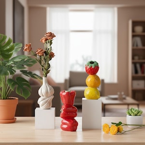Peut inclure: Trois vases en céramique en forme d'aliments : un vase bulbe d'ail blanc contenant des roses séchées, un vase empilé en forme de poivron rouge et un vase empilé en forme d'agrumes jaunes surmonté d'une fraise, posés sur des socles blancs.