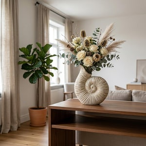 Puede incluir: Un jarrón de cerámica color crema con forma de concha de nautilo, lleno de un arreglo floral de rosas blancas, hierba de la pampa seca, hojas de eucalipto y vainas de semillas marrones, creando un centro de mesa decorativo y texturizado.