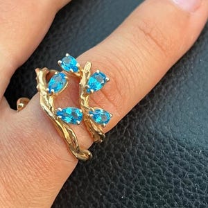 Può includere: Un anello in oro con un design a fascia testurizzata simile a un ramo, decorato con cinque pietre preziose blu brillante a forma di pera. Il metallo ha una finitura opaca e organica, e le pietre sono disposte a grappolo sulla parte superiore.