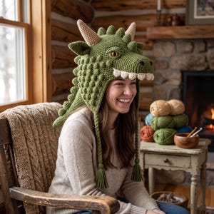 May include: A green crocheted dragon hat featuring a textured scale pattern, two light-colored horns, a row of white teeth along the front edge, and two long braided earflaps with tassels at the ends. The hat has a prominent snout and eyes.