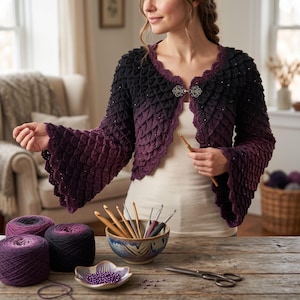 May include: A crochet cardigan featuring a textured scale pattern that transitions from black at the shoulders to deep purple at the flared sleeves. The garment has a scalloped edge, a silver-toned ornate metal clasp at the neckline, and small beads.