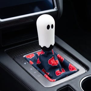 May include: A white ghost-shaped gear shift knob with black oval eyes sits atop a dark blue fabric shift boot featuring a repeating pattern of red clouds. The gear shifter is installed in a vehicle center console with a silver trim and black interior.