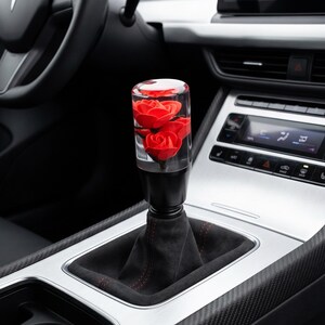 May include: A cylindrical gear shift knob featuring a clear acrylic top encasing three vibrant red roses. The base of the knob is solid black, mounted onto a black fabric gear shift boot with subtle red stitching, positioned in a vehicle console.