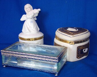 May include: Three decorative boxes, one with a porcelain angel figurine playing a musical instrument, one with a white swan design, and one with a clear glass top and silver trim.
