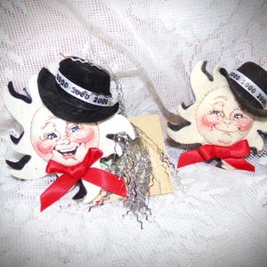 May include: Two whimsical sun-shaped ornaments with smiling faces, rosy cheeks, and red bow ties. Each sun wears a small black hat with the year "2000" on the band. The ornaments have black accents and decorative fringe.