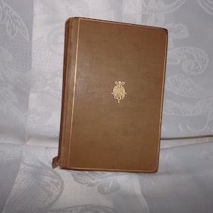 May include: A vintage book with a tan cover and gold trim. A gold emblem is centered on the front. The book's spine is visible, and the pages are slightly aged. The book is standing upright.