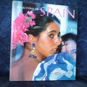May include: A book cover titled "Reflections of Spain" featuring a woman with pink flowers in her hair and blue earrings. The word "SPAIN" is in pink letters. The woman is wearing a blue and white shawl.