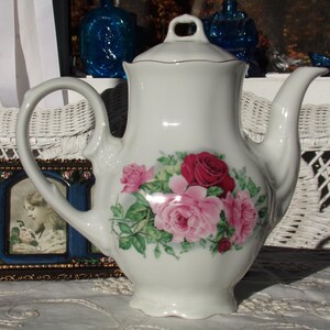 May include: White porcelain teapot with a floral design featuring pink and red roses. The teapot has a classic shape with a curved spout and a lid with a knob.