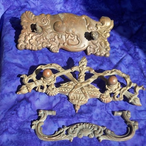 May include: Three antique brass drawer pulls with intricate floral designs. The pulls are ornate and feature a variety of shapes and curves.