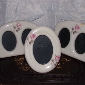 May include: Three white ceramic picture frames with oval openings. Each frame features a pink rose with green leaves painted on the front.