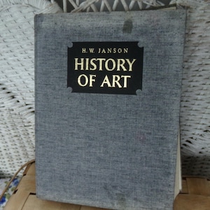 May include: A hardcover book titled "HISTORY OF ART" by H.W. Janson. The book has a gray cover with the title in gold lettering on a black label. The book is standing upright against a white wicker background.