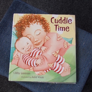 May include: A children's book titled "Cuddle Time" featuring an illustration of a mother and baby. The baby is wearing a red and white striped outfit. The book is on a blue cloth.