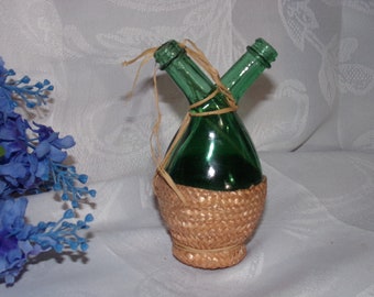 May include: A green glass cruet with two spouts, wrapped with a woven straw basket. The cruet is tied with raffia and is designed for dispensing oil and vinegar. The background is a white fabric with a blue floral arrangement.