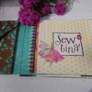 May include: A brown fabric-covered journal with a teal and white patterned spine. The front cover is yellow with white polka dots and a pink fairy with wings. The title "Sew tina!" is written in brown cursive lettering.