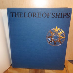 May include: A blue book titled "THE LORE OF SHIPS" with gold lettering and a gold compass rose design. The book is hardcover and appears to be in good condition. The book is on a wooden surface.