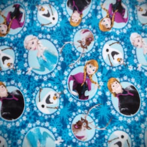May include: A blue fabric featuring a repeating pattern of characters from the animated film "Frozen." The design includes images of Elsa, Anna, Olaf, and Sven, set against a backdrop of snowflakes and circular frames.