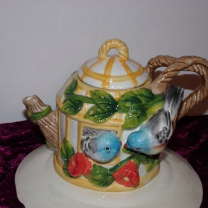 May include: A white ceramic teapot shaped like a birdcage with two blue birds perched on the side. The teapot has a yellow and white striped lid and a brown rope handle. The teapot is sitting on a white saucer.