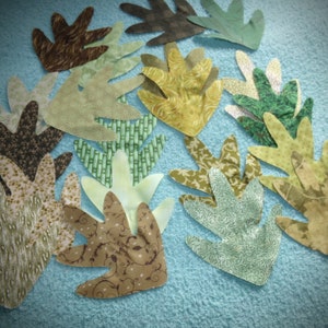 May include: A collection of fabric leaves in various shades of green, brown, and yellow. The leaves are cut in a simple, hand-crafted style and have a variety of patterns.