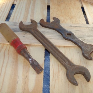 May include: Three rusty tools on a wooden surface. A red-handled chisel, a large wrench, and a smaller wrench with the text "Williams  Wrench Co.  Wrenches" on the handle.