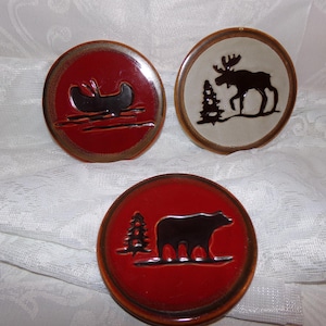May include: Set of three red and white ceramic coasters with black silhouette designs. One coaster features a canoe, another a moose, and the last a bear.
